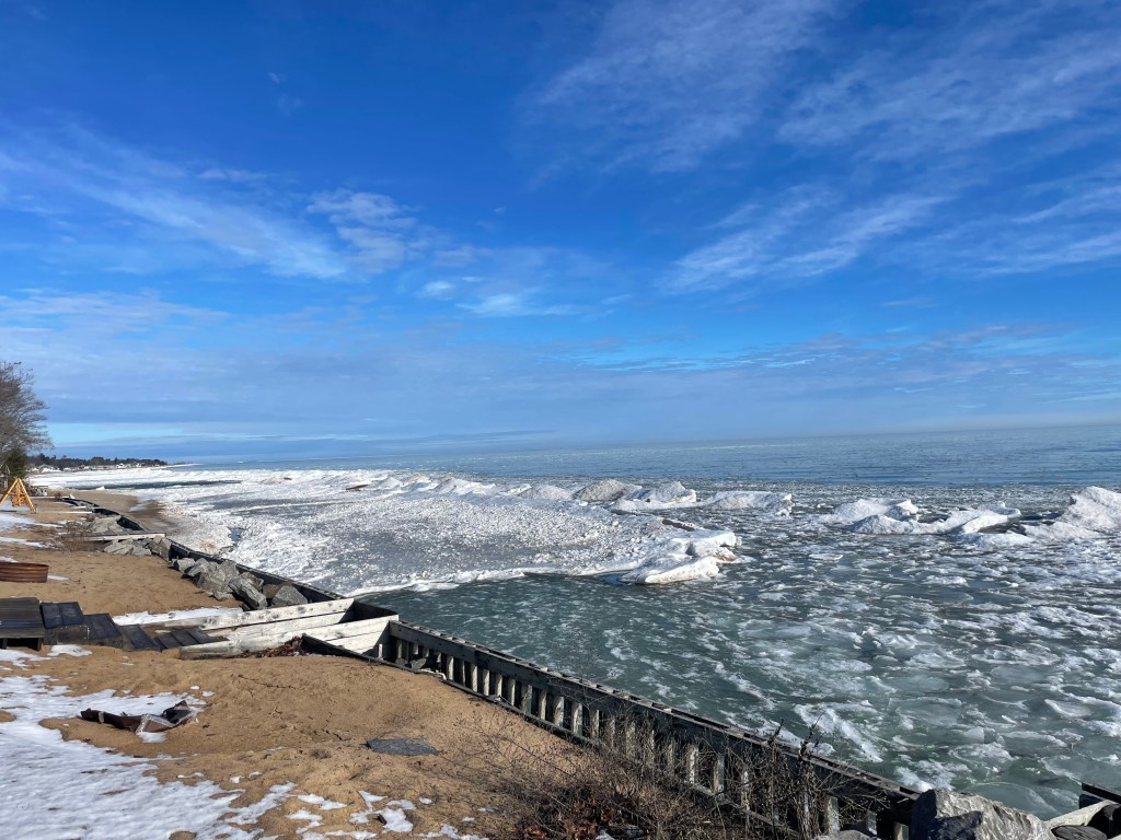 An ice shelf and pieces of floating ice line a residential seawall on Lake Huron on a sunny day.